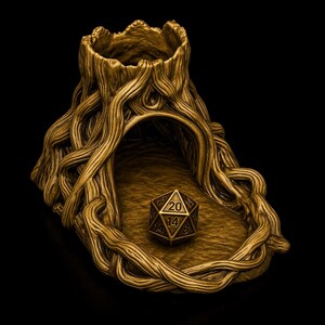 May include: A metallic gold-colored dice tower and tray set featuring a sculpted tree stump design with intricate root details. A single twenty-sided die with black numbers is positioned in the center of the tray area in front of the tower.