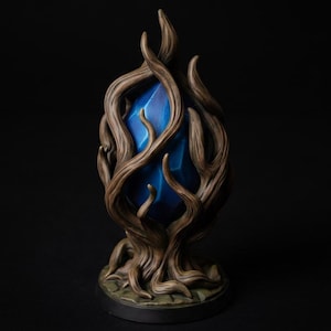 Puede incluir: Una escultura decorativa que presenta un cristal azul brillante y facetado encerrado en una estructura marrón nudosa similar a una raíz. La base es circular y oscura, sosteniendo las ramas entrelazadas de textura leñosa que rodean la gema.