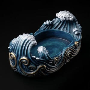 May include: An oval-shaped decorative bowl featuring a sculpted wave design. The exterior and interior are finished in deep blue tones with white highlights on the crests of the waves and metallic gold accents along the swirling base.