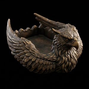Puede incluir: Un cuenco decorativo de color bronce con forma de grifo mítico, con una cabeza de águila detallada y alas emplumadas que se extienden por los lados. El cuenco central es circular y liso, rodeado por el cuerpo esculpido de la criatura.
