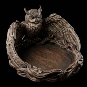 May include: A brown, carved bowl featuring a detailed owl with spread wings forming the sides of the vessel. The base of the bowl is shaped like intertwined branches and leaves, creating a circular, open center for storage or display.