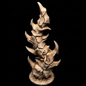 Op de afbeelding: Een verticale, vlamvormige sculptuur met een getextureerd, botkleurig oppervlak met donkerbruine verwering in de groeven. Het object heeft een gelaagd, grillig uiterlijk met meerdere puntige uitsteeksels die uit een centrale basis komen.