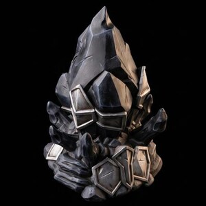 Op de afbeelding: Een sculpturaal object dat lijkt op een cluster van donkere, grillige kristallen of rotsformaties. De structuur heeft scherpe, hoekige facetten in tinten houtskool en zwart, geaccentueerd met metallic zilverkleurige geometrische platen.