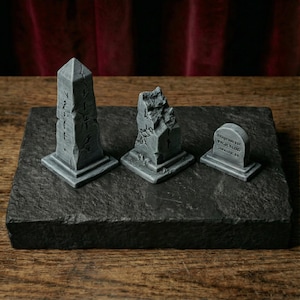 Peut inclure: Trois monuments miniatures en pierre grise comprenant un obélisque haut avec des marques gravées, un pilier brisé et déchiqueté, et une petite pierre tombale arrondie. Chaque pièce présente une surface texturée et vieillie.