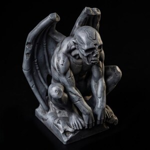 May include: A grey, weathered gargoyle statue in a crouching position with large, folded wings. The figure features a textured, stone-like surface with visible cracks and crevices across its muscular body, head, and wings, set against a dark background.