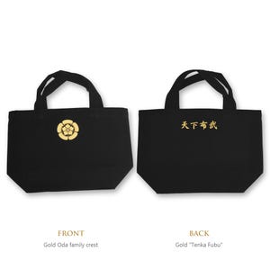 Puede incluir: Un bolso tote negro con dos asas. La parte delantera presenta un escudo familiar Oda dorado. La parte trasera muestra el texto japonés dorado "天下布武". El bolso tiene una base rectangular y una abertura amplia, con un diseño sencillo.