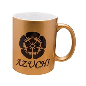 Peut inclure: Une tasse en céramique dorée métallisée avec un intérieur blanc et une anse incurvée. L'avant présente un emblème floral circulaire noir au-dessus du texte AZUCHI imprimé dans une police serif foncée. La tasse a une finition lisse.