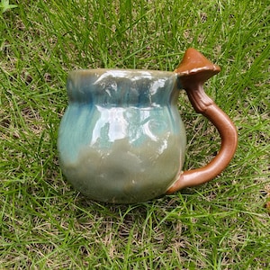 Puede incluir: Una taza de cerámica con cuerpo redondeado y un esmalte brillante que transiciona de un azul profundo en el borde a un verde salvia apagado en la base. El asa está esculpida con la forma de un hongo marrón con sombrero y tallo.