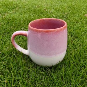 May include: A ceramic mug featuring a two-tone design with a soft pink upper section and a solid white base. The rim and the curved handle are finished in a slightly darker, muted coral-pink glaze that contrasts with the lighter pink body.