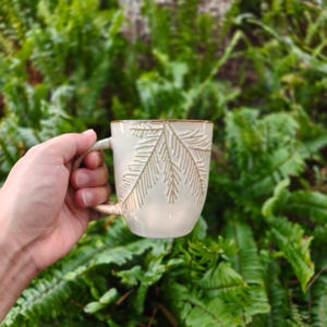 May include: A cream-coloured ceramic mug featuring a textured, debossed fern leaf pattern on the exterior. The mug has a smooth, glossy finish and a curved handle on the side, showcasing a natural, earthy aesthetic with detailed botanical carvings.