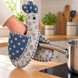 May include: A chicken-shaped oven mitt featuring a light gray body with a blue floral pattern, a dark blue floral crest, and a mustard yellow beak. The mitt has a blue and white checkered trim and a dark blue floral wing-shaped pocket.