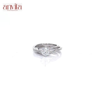 Peut inclure: Une bague argentée avec une pierre centrale ronde montée sur quatre griffes, accompagnée de petites pierres incrustées le long de l'anneau divisé. Le nom de marque anvita est imprimé en rouge dans le coin supérieur gauche de l'image.