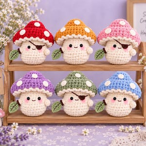 Puede incluir: Seis pequeñas figuras de hongos tejidas a ganchillo, redondas, con cuerpos de color crema y sombreros coloridos en rojo, naranja, rosa, morado, verde y azul. Cada hongo presenta puntos blancos, una cara sonriente, mejillas rosadas y una hoja verde.
