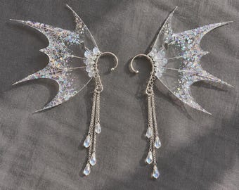 May include: A pair of silver-toned ear cuffs featuring translucent, wing-shaped attachments filled with iridescent glitter. Each cuff includes a cluster of small clear beads and three delicate silver chains ending in teardrop-shaped charms.