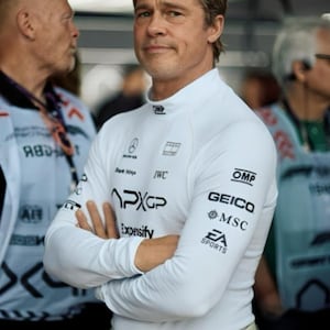May include: A white long-sleeve racing shirt with a mock neck collar, featuring various black sponsor logos including APXGP, OMP, GEICO, MSC, and EA Sports printed on the chest and sleeves, worn with light-colored racing trousers at the waist.