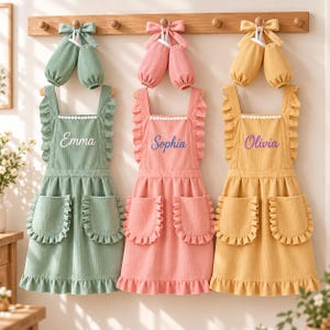 May include: Three corduroy aprons in sage green, dusty pink, and mustard yellow, each with matching arm sleeves and a bow. The aprons feature ruffled straps, two ruffled pockets, and white lace trim. The names Emma, Sophia, and Olivia are embroidered.