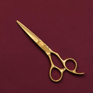May include: A pair of gold-colored metal hair cutting shears featuring a polished finish, two finger rings, and a curved finger rest. The blades are straight and pointed, with a screw mechanism connecting the two halves of the tool.