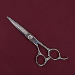 Puede incluir: Tijeras de corte de pelo de acero inoxidable de color plateado con dos anillos para los dedos, un tornillo central de ajuste de tensión y un apoyo para el dedo curvo que se extiende desde uno de los mangos. Las hojas son rectas y puntiagudas.