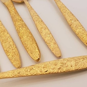 May include: Close-up of a set of gold-colored flatware, including forks and spoons. The handles are intricately decorated with a floral pattern. The flatware is arranged on a white surface.