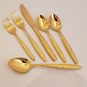 May include: A set of six gold-colored flatware pieces, including forks, knives, and spoons. The utensils have textured handles and a shiny, reflective surface. The arrangement suggests a formal dining setting.