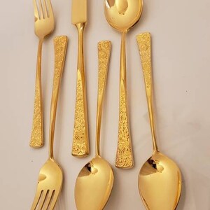 May include: A set of gold-colored flatware, including a knife, fork, and spoons. The flatware has an ornate design.