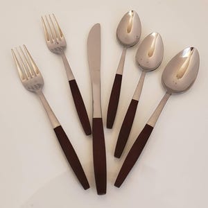 May include: A set of stainless steel silverware with dark brown wooden handles. The set includes two forks, a knife, and three spoons. The silverware is arranged on a white surface.
