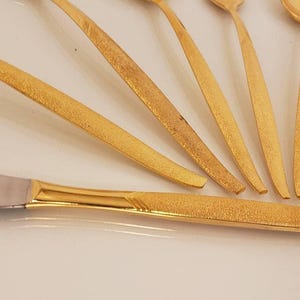 May include: A collection of gold-colored flatware, including spoons and a knife, arranged on a white surface. The knife has a silver blade and a textured gold handle. The spoons have a similar textured gold finish.