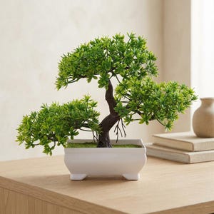 Könnte beinhalten: Ein kleiner künstlicher Bonsai-Baum mit leuchtend grünem Blattwerk und einem dunkelbraunen, strukturierten Stamm. Der Baum ist in einen rechteckigen, weißen Keramiktopf mit vier kleinen Füßen gepflanzt und hat eine moosartige grüne Basis.