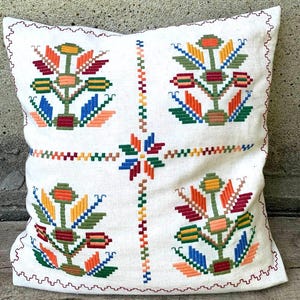 May include: A square off-white throw pillow featuring a cross-stitch embroidery pattern of four symmetrical floral motifs in vibrant shades of orange, yellow, green, blue, and red, with a central star design and a decorative zigzag border.