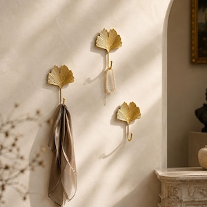 May include: Three gold-colored wall hooks shaped like ginkgo leaves. One hook holds a brown scarf, another holds a white beaded necklace, and the third hook is empty. The hooks are mounted on a light-colored wall with soft shadows cast across them.