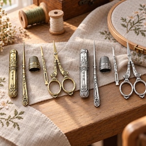 Könnte beinhalten: Zwei Sets von Nähwerkzeugen, eines in goldfarbenem und eines in silberfarbenem Metall. Jedes Set enthält ein Nadelgehäuse, eine lange Nadel, einen Fingerhut und verzierte Scheren. Die Werkzeuge weisen detaillierte florale Gravuren auf.
