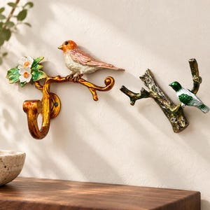 Puede incluir: Dos ganchos de pared con forma de ramas de árbol. El gancho izquierdo presenta una rama dorada con un pájaro naranja posado y flores blancas con hojas verdes. El gancho derecho es una rama oscura texturizada con un pequeño pájaro verde y blanco.