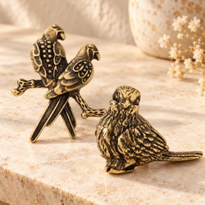 May include: Two antique brass-toned metal bird figurines. One piece features two birds perched together on a branch with detailed wing textures. The second piece is a single bird figurine with textured plumage, facing forward with a long tail.