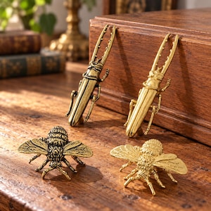 May include: A collection of four metal cabinet knobs featuring insect designs. Two knobs are shaped like elongated stag beetles with gold-toned finishes, and two knobs are shaped like bees with textured wings and bodies in gold-toned metal.