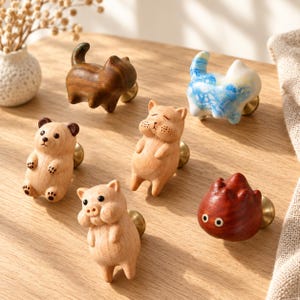 Op de afbeelding: Een verzameling van zes decoratieve kastknoppen in de vorm van dieren, waaronder een donkerbruine houten kat, een blauw met wit gemarmerde kat, een lichthouten beer, een lichthouten hamster, een lichthouten varken en een donkerrood houten wezen.