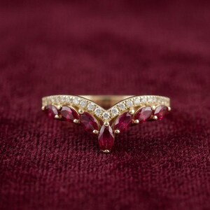 Op de afbeelding: Een V-vormige gouden ring met een rij rode robijnen in marquise-slijpvorm langs de onderrand en een rij kleine ronde witte diamanten langs de bovenrand, wat een chevron-ontwerp vormt dat de contouren van de edelstenen volgt.