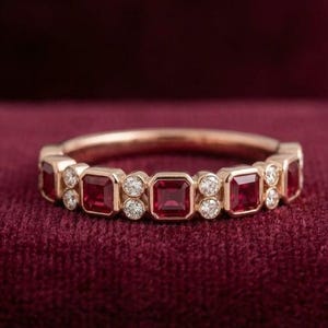 May include: A rose gold band ring featuring an alternating pattern of square-cut red rubies and small round white diamonds. The gemstones are set in a continuous row around the band, creating a symmetrical design with a polished metal finish.