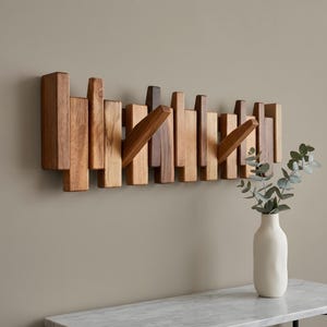 May include: A wall-mounted coat rack constructed from multiple vertical wooden slats of varying lengths and brown tones. The rack features two angled wooden pegs extending outward from the slat arrangement to provide functional hanging points.