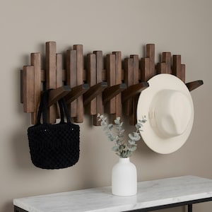 May include: A wall-mounted coat rack made of dark brown wood with vertical slats of varying heights and five protruding hooks. A black crocheted handbag hangs from one hook, and a cream-colored wide-brimmed hat hangs from another hook.