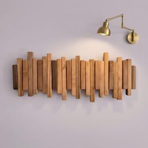 May include: A wall-mounted coat rack composed of numerous vertical wooden slats of varying lengths and shades of brown, arranged in a staggered, overlapping pattern. Above the rack, a brass-colored articulated wall lamp is mounted.