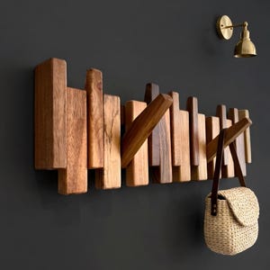 May include: A wall-mounted coat rack constructed from vertical wooden slats of varying lengths and shades of brown. Two angled wooden pegs extend outward from the rack, with a woven straw handbag featuring a dark brown leather strap hanging from one.