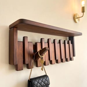 May include: A wall-mounted dark wood shelf featuring a series of vertical wooden slats of varying lengths. A single wooden hook extends from the center, holding a gold-toned key ring with a black quilted bag attached by a gold chain.
