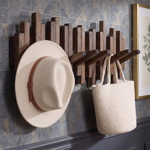 May include: A wall-mounted coat rack constructed from vertical dark brown wooden slats of varying heights, featuring several protruding angled hooks. A cream-colored felt hat with a brown leather band and a cream woven tote bag hang from the hooks.
