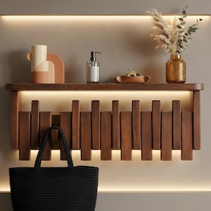 May include: A wall-mounted wooden shelf featuring a series of vertical slats and integrated LED lighting. The shelf holds a geometric vase, a clear soap dispenser, a small leather tray with keys, and a textured amber glass vase with dried foliage.