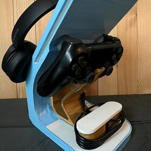 May include: A light blue gaming stand featuring a wooden support structure, holding a black game controller and black headphones. A white base component sits at the front with a coiled black charging cable resting on top of the flat surface.