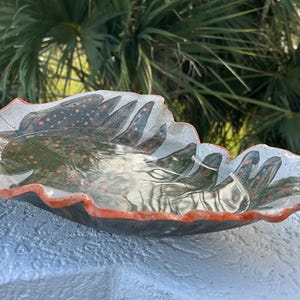 May include: A ceramic leaf-shaped bowl with a dark green and red speckled design. The rim is outlined in orange. The bowl is sitting on a white surface, with a blurred background of green palm fronds.