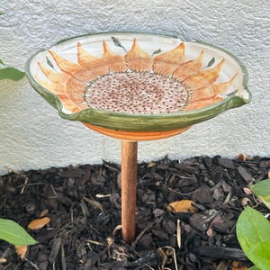 May include: A ceramic bird bath with a sunflower design. The bowl is painted with orange and brown petals and a brown center. The rim is green and orange. It is mounted on a copper-colored metal pole, set in dark mulch.