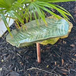 May include: A green ceramic bird bath with a leaf design, supported by a copper rod and a stone base. The bird bath is surrounded by black mulch and green palm fronds.