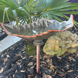 May include: A ceramic bird bath with a leaf-shaped design. The bath has a gray interior with a red-orange rim and is supported by a copper-colored metal stake. The bird bath is filled with water and sits in a garden bed with mulch and foliage.