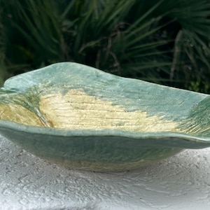 May include: A handcrafted, ceramic serving bowl with a leaf-like design. The bowl features a light green exterior and a golden interior, with a textured surface. The bowl is displayed outdoors, against a backdrop of green foliage.